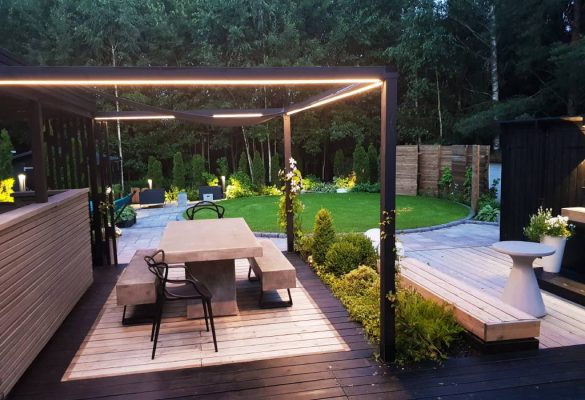 Kontio-piha-4-1-1 - LedStore Moderni takapihan patio, jossa on betoninen ruokapöytä ja mustat tuolit minimalistisen pergolan alla. Pergolaan on integroitu LED-valaistus luomaan tunnelmallista ilmettä. Aluetta ympäröivehreä kasvillisuus, yksi hoidettu nurmikko ja kivipolku, joka lisää luonnonläheistä viehätystä. Lisäksi pienenä yksityiskohtana löytyy valkoinen moderni istutuslaatikko elegantilla terassilla nauttimista varten. Tutustu LedStore.fi:n laajaan LED-valaistusvalikoimaan ja tuo samaa modernia eleganssia omaan pihaasi!