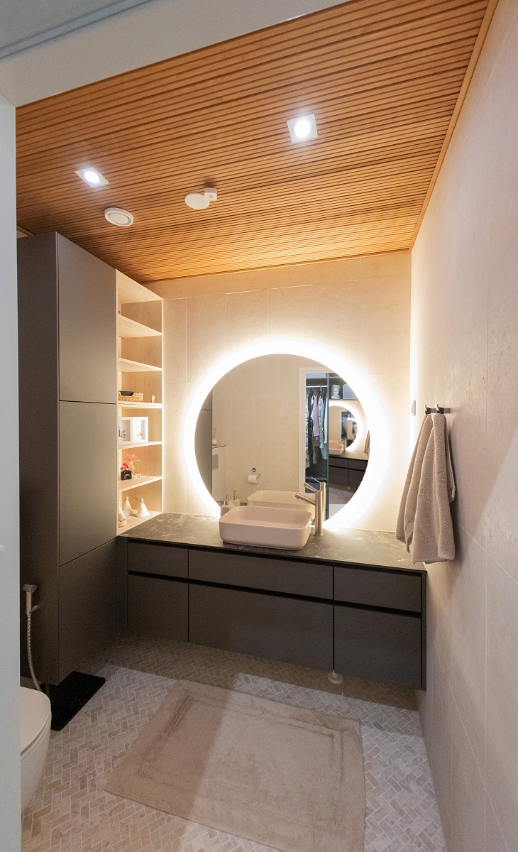 LedStore.fi-verkkokaupan LED-valaistusta hyödyntävä moderni kylpyhuone: Pesualtaan yläpuolella on pyöreä LED-taustavalaistu peili, joka luo tilaan eleganttia tunnelmaa. Kylpyhuoneessa on tummat kaapit, lämminhenkinen puukatot sekä vaaleat seinät. Oikealle puolesta on aseteltu pyyhkeet ja vasemmalla odottaat hyllyt. Lattialla lepää beige matto, joka osaae kokonaisuuden.