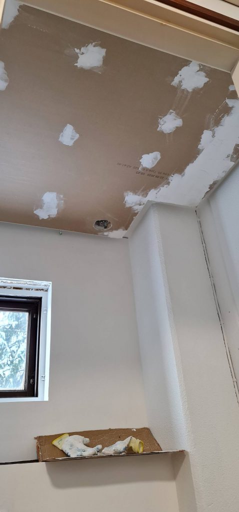 Katon saneeraus kipsilevypaikoilla, luonnonvalo avoimesta ikkunasta vasemmalla, hienovarainen LED-nauhavalaistus, pahvi ja hanskat kielekkeellä.