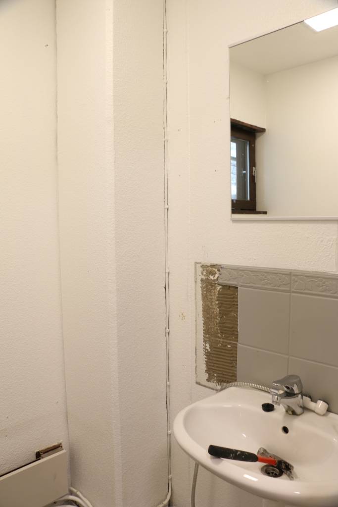 Pieni kylpyhuone, jossa LED-kehystetty peili valkoisen pesualtaan yläpuolella, hopea hana, esillä oleva kaapeli; musta ja punainen jakoavain pesuallas. Osittain poistetut laatat paljastavat seinän.