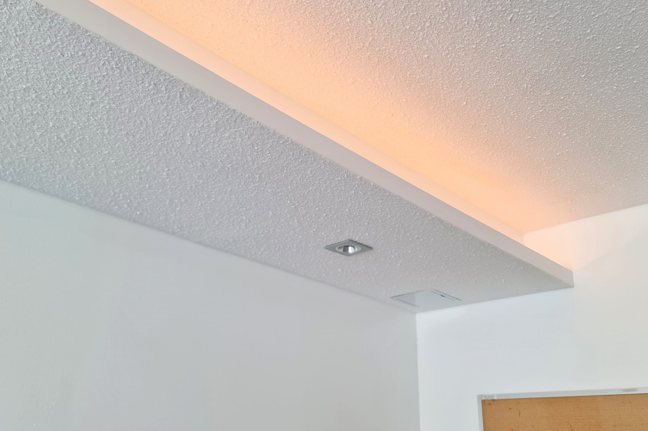 Hyllyn päällä valmis led-nauha - LedStore Sisäkaton nurkka, johon on upotettu LED-valaistus ja jossa on teksturoitu valkoinen pinta. Keskellä kattoa näkyy pieni, neliönmuotoinen valaisin. Valaistus luo lämpimän oranssihehkun katon reunaa pitkin.
