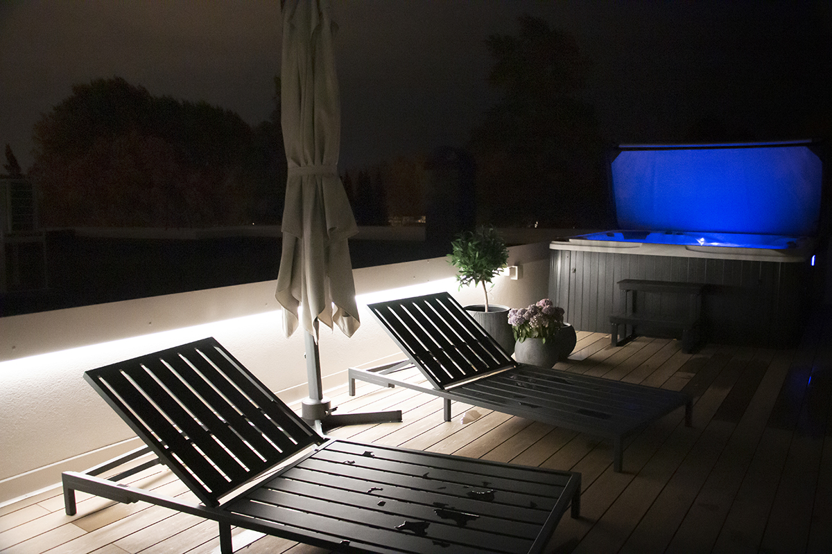 Kaksi lepotuolia ja suljettu aurinkovarjo terassilla illan hämärässä. LedStore.fi:n laadukas led-pihavalaistus valaisee pehmeästi taustalla hehkuvaa, sinisävyistä poreallasta tämän modernin talon pihapiirissä.