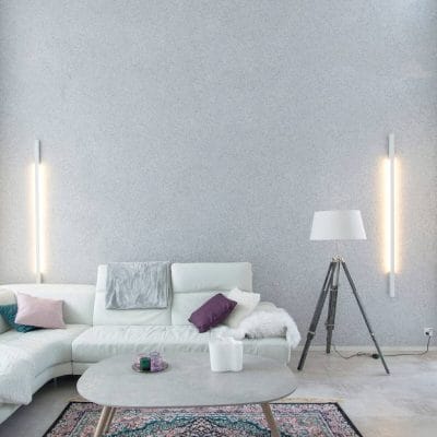 Moderni olohuone, jossa L-muotoinen sohva, vaaleanpunaiset/violetit tyynyt, pyöreä sohvapöytä, LED-lattiavalaisin, seinävalaisimet, koristeellinen matto kuvioitua harmaata seinää vasten.