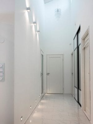 Minimalistinen valkoinen käytävä LED-seinävalaisimilla, laattalattialla ja kattokruunulla. Suljettu ovi päässä ja sivuovet lisäävät kirkkautta.