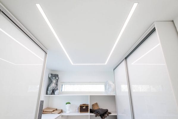 Moderni minimalistinen vaatekaappi valkoisilla hyllyillä, liukuovilla, koriste-esineillä, koreilla, taitetuilla peitoilla. Valaistu upotettu LED-valaistus ja ikkuna.