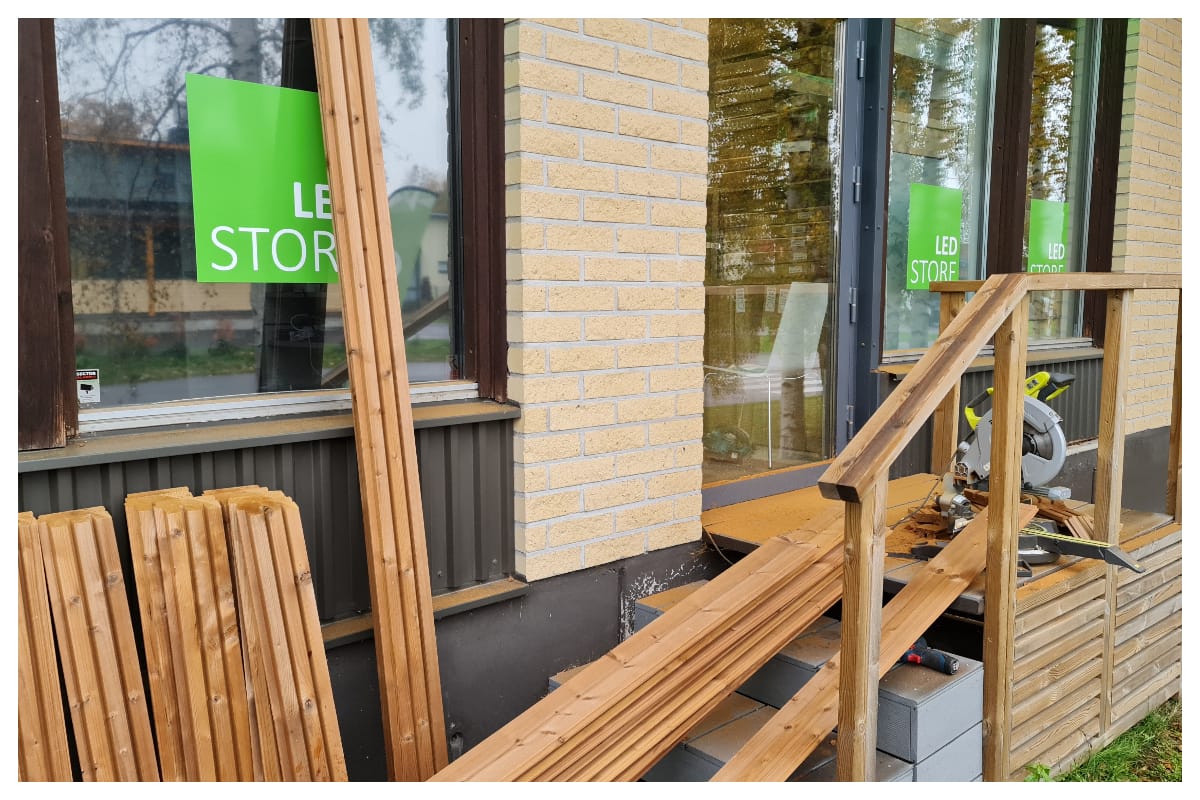 LedStore.fi-verkkokaupan muuten, osittain rakennetun rampin viereen on aseteltu pinoja puulankkuja. Tiilirakennuksen ikkunoissa loistavat vihreät kyltit julistavat kirkkaasti "LED STORE." Saha lepää rampilla, valmiina toimintaan.