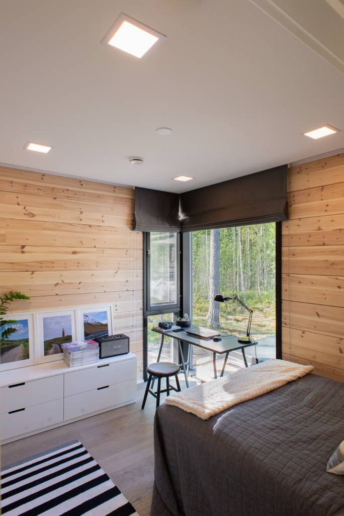 - LedStore Viihtyisä puuseinäinen huone, jossa on LED-pöytävalaisin, yhden hengen sänky, kirjoituspöytä metsänäköalan ikkunoiden vieressä, musta tuoli, kirjoja, kehystetty taide, kasvi lipaston päällä.