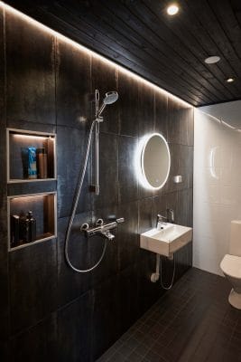 Tyylikäs kylpyhuone tummat laatat, LED-taustavalaistu peili pesualtaan yläpuolella, suihku, sisäänrakennetut hyllyt ja musta katto. Moderni wc täydentää tyylikkään ilmeen.