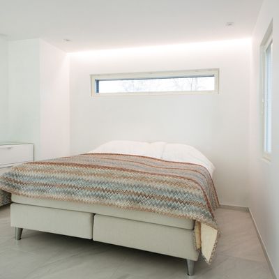 Ein minimalistisches Schlafzimmer mit Kingsize-Bett, gemusterter Tagesdecke, kleinem Fenster über dem Kopfteil und heller LED-Beleuchtung in einem weißen Raum.