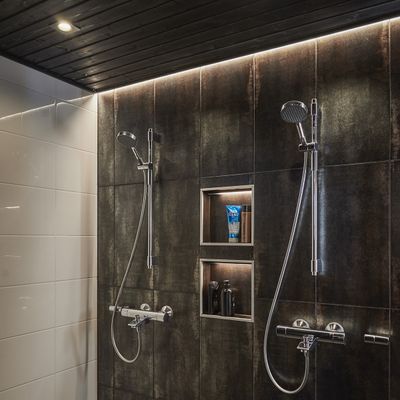 Moderne Dusche mit zwei Duschköpfen, dunkel gefliester Wand, eingebauten Regalen mit Toilettenartikeln und hinterleuchteter Holzdecke für eine elegante Atmosphäre.