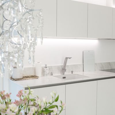 Moderne weiße Küche mit Arbeitsflächen aus Marmor, LED-Unterschrankbeleuchtung, Kristalllüster und rosa Blumendekoration im Vordergrund.