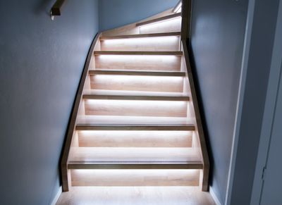 Eine moderne Holztreppe mit LED-Leuchten unter jeder Stufe, die das minimalistische Design mit warmer Beleuchtung vor grauen Wänden hervorheben.