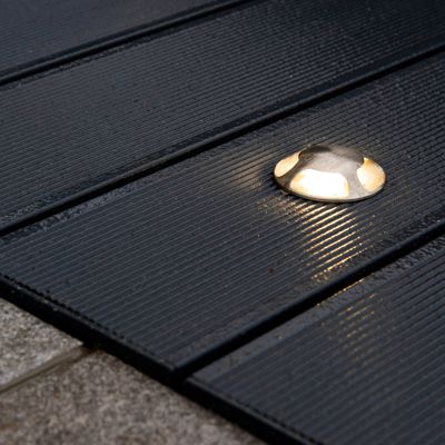 Silberfarbene, runde Terrassenleuchte mit dunklen, gerillten Holzbrettern, die nachts ein sanftes Licht abgibt. Ideal für LED-Beleuchtungslösungen im Außenbereich.