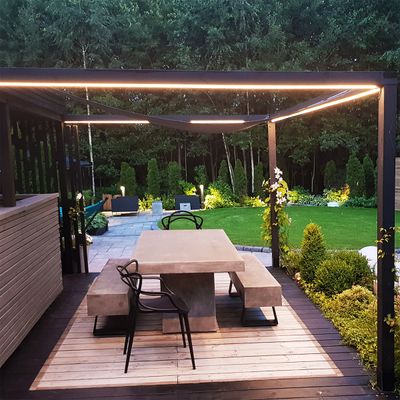 Moderne Terrasse mit Steintisch und Bänken, Pergola mit LED-Beleuchtung, beschnittenen Sträuchern und üppigem Rasen. Ein perfektes Beispiel für LED-Außenbeleuchtung.