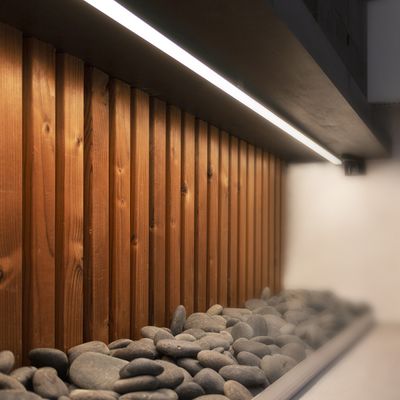 Wand aus Holzpaneelen mit vertikalen Linien, beleuchtet von LED-Leuchtbändern auf glatten grauen Steinen für ein modernes Ambiente.