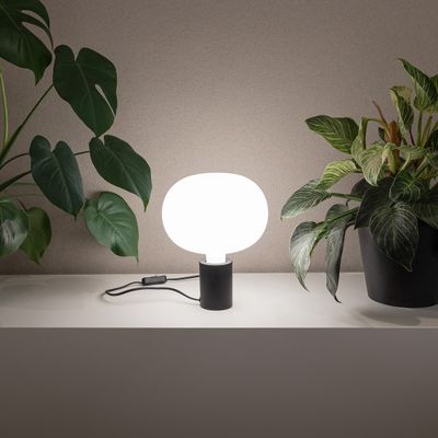 Moderne LED-Tischleuchte mit weißem, rundem Schirm auf einem Regal, umgeben von grünen Topfpflanzen für eine elegante Beleuchtung zu Hause.