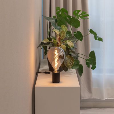 Vintage-LED-Lampe, die einen weißen Tisch neben einer großen grünen Pflanze vor einer beigen Wand mit transparenten Vorhängen im Hintergrund beleuchtet.