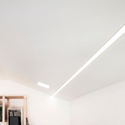 Minimalistischer weißer Raum mit schräger Decke und linearer LED-Einbauleuchte mit einem kleinen weißen Regal mit verschiedenen Gegenständen in einer Ecke.