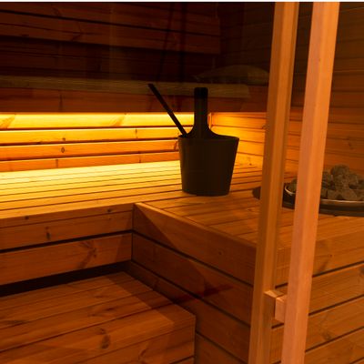 Inneneinrichtung der Holzsauna mit Bänken, sanfter LED-Beleuchtung, schwarzem Eimer und Schaufel auf der Bank für eine gemütliche und entspannende Atmosphäre.