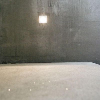 Ein minimalistischer Raum mit grauen Wänden und Boden, mit einer kleinen quadratischen LED-Leuchte an der Rückwand.