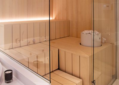 Moderne Saunakabine mit stilvollen hellen Holzbänken, Glaswänden und einem Eimer mit weißen Steinen neben der LED-Beleuchtung neben dem Ofen.