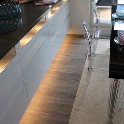 Moderne Küche mit glänzenden Schränken, LED-Beleuchtung unter dem Schrank, transparentem Stuhl, schwarzen Arbeitsplatten und stilvollem Holzboden.