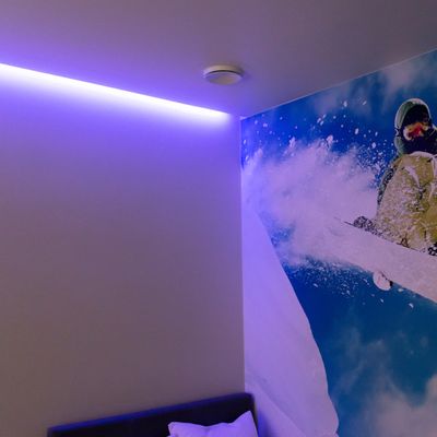 Zimmer mit weißen Wänden, lila LED-Deckenleuchte, Skifahrer-Gemälde an der Wand; sichtbares Kissen und Teil des Bettes. Moderne und gemütliche Beleuchtungslösung für das Schlafzimmer.