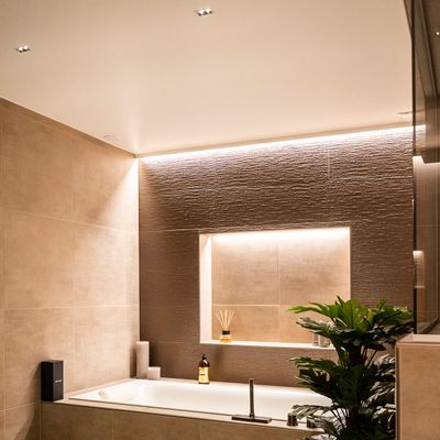 Modernes Badezimmer mit Badewanne, LED-Einbauleuchten, beigen Fliesen, Topfpflanze; Regale mit Kerzen und Toilettenartikeln.