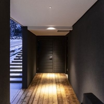 Alternativer Text: Moderne Eingangshalle mit dunklen Wänden, LED-beleuchtetem Holzfußboden und Treppe; schwarze Tür, die nach draußen in den Schnee führt. Perfekte Atmosphäre für elegante Innenräume.