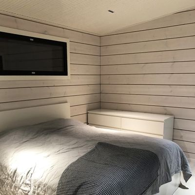 Ein gemütliches Schlafzimmer mit holzgetäfelten Wänden, stimmungsvoller LED-Beleuchtung, grauer Bettwäsche auf dem Bett und einer weißen Kommode unter einem kleinen Fenster, um eine friedliche Atmosphäre zu schaffen.