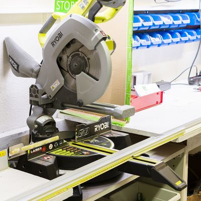 Alternativer Text: eine Ryobi Gehrungssäge auf einer Werkbank, umgeben von geordneten Regalen mit kleinen blauen Behältern und verschiedenen Werkzeugen in einer gut beleuchteten LED-Beleuchtungslösung.
