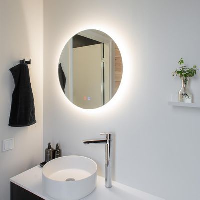 Minimalistisches Badezimmer mit rundem Spiegel mit LED-Beleuchtung, weißem Waschbecken mit verchromtem Wasserhahn, schwarzem Handtuch und kleiner Pflanze auf dem Regal.