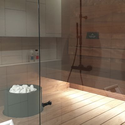 Moderne Saunakabine mit Holzwänden, Glastür, Steinkübel, Duschkopf; perfekte Atmosphäre mit LED-Beleuchtung für Entspannung und Komfort.