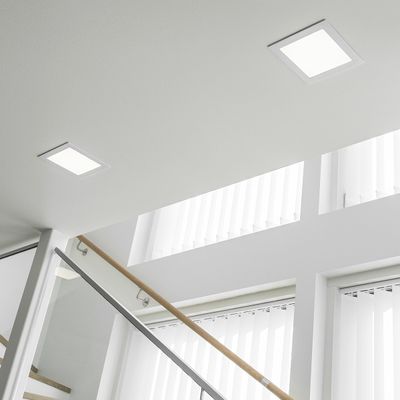 Helle quadratische Deckeneinbauleuchten beleuchten ein modernes Treppenhaus mit Glasgeländer und großen Fenstern mit vertikalen Vorhängen.
