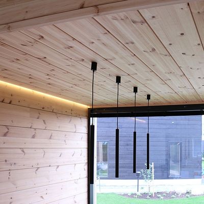 Ein modernes Interieur mit Holzwänden und -decken, vier schwarzen Pendelleuchten und einem großen Fenster zum Garten hin.