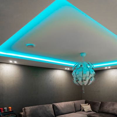 Ein modernes Wohnzimmer mit einem grauen Sofa, blaugrünen LED-Deckenleuchten, einer runden Pendelleuchte und einem kleinen Tisch mit lebhaften Kerzen für die Raumbeleuchtung.