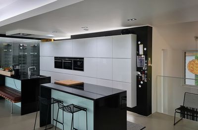 Moderne Küche mit glänzend weißen Schränken, schwarzen Arbeitsplatten, Insel mit Barhockern, Doppel-Backofen aus Edelstahl, Kühlschrank mit Magneten direkt unter LED-Beleuchtung.