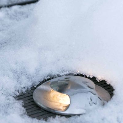 Alternativer Text: "Teilweise schneebedecktes Bodenlicht, das einen warmen Schein in die Winterlandschaft zaubert - ein Produktschaufenster für das LED-Beleuchtungsunternehmen.