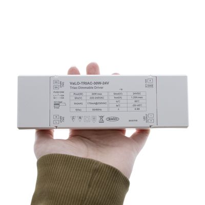 Eine Person mit dem "VaLO-TRIAC-30W-24V" LED-Regler mit Angaben auf einer weißen Oberfläche, ideal für LED-Beleuchtungssysteme.