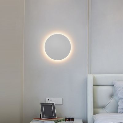 Eine moderne runde Wandleuchte beleuchtet ein minimalistisches Schlafzimmer mit hellgrauen Wänden, einem weißen getufteten Bettkopfteil und einer stilvollen Nachttischdekoration. LED-Beleuchtung Shop.