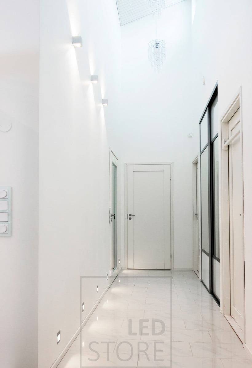CUBIC LED-Wandleuchte, die in einem Korridor in zwei Richtungen leuchtet, um die Höhe des Raums zu betonen. Eingebaute LED-Wandleuchten am Bodenrand sorgen für indirektes Licht auf dem Boden. Ledstore.fi