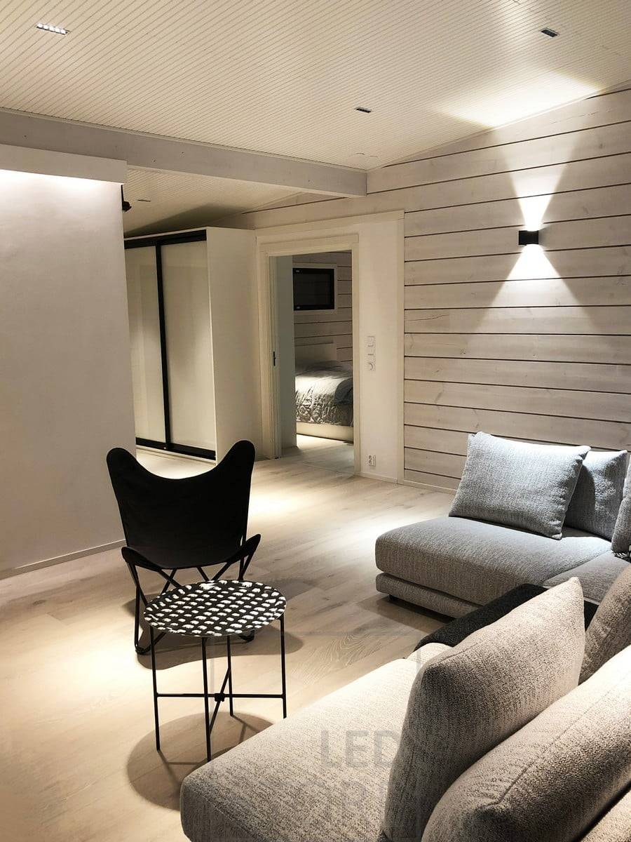 Schwarze FUNK-Wandleuchte als Teil der stimmungsvollen Beleuchtung des Wohnzimmers. Neben der FUNK-Wandleuchte wird der Raum durch LINJA2-Spots an der Decke und einen LED-Streifen hinter der Leiste beleuchtet. Ledstore.fi