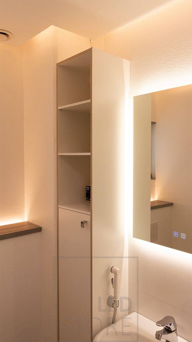 Sanfte Beleuchtung in einem Badezimmer mit vollständig indirektem Licht. Led-Lichtleisten hinter der Toilettenkabine, die nach oben gerichtet sind, und an der Decke in einem Kasten, der die Wand nach unten beleuchtet, das Ganze gekrönt von einem Lichtspiegel. Ledstore.fi