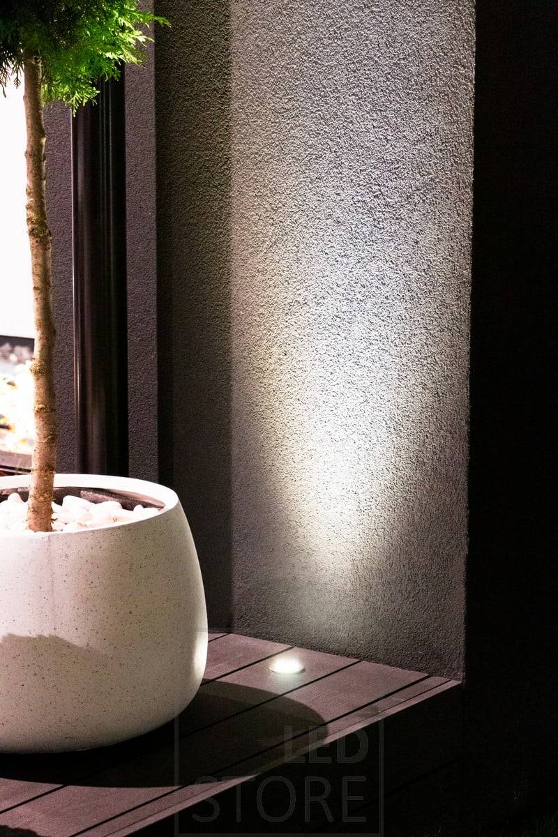 In die Terrasse eingelassener, nach oben strahlender Strahler, der durch die Wand hindurch leuchtet. Das Licht hebt auch die Pflanze im Vordergrund schön hervor. Ledstore.fi