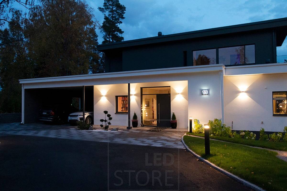 Weiße bidirektionale FUNK-Wandleuchte, die die Fassade und den Eingang beleuchtet. Die Einfahrt wird von Säulenleuchten beleuchtet. Ledstore.fi