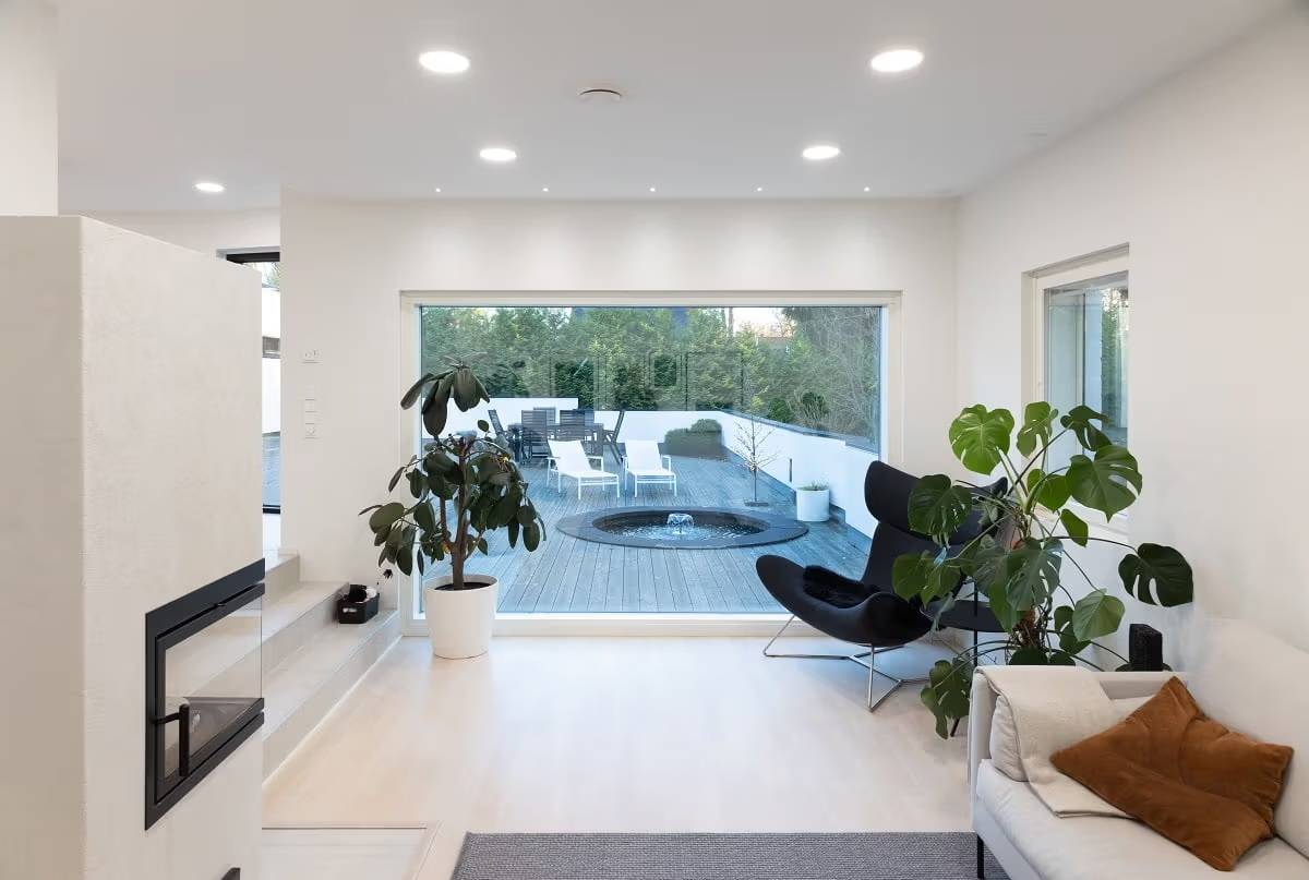 Ein Wohnzimmer mit einer hellen Oberfläche und einem großen Fenster mit Blick nach draußen. An der Decke befinden sich runde LED-Paneele und vor dem Fenster kleine Strahler.