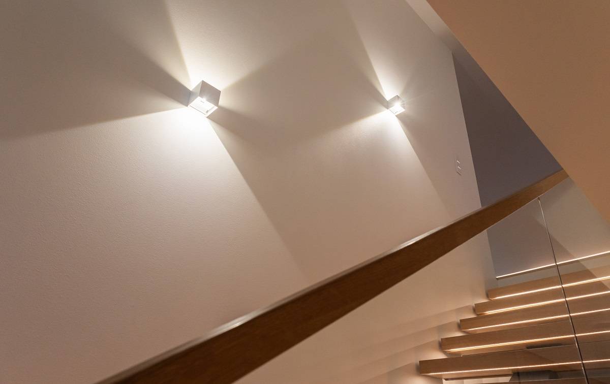 Eine moderne Treppe mit Holzstufen und Glasgeländer erhält einen tollen Look mit den stilvollen LED-Wandleuchten von LedStore.fi, die schöne Lichtmuster an der Wand erzeugen. Wählen Sie eine Designer-Beleuchtung, die Ihrem Zuhause einen modernen Look verleiht!