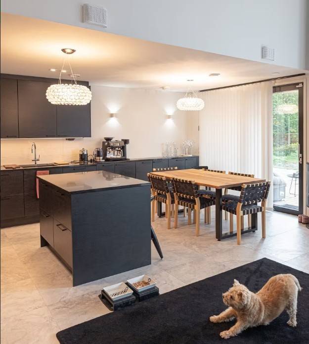 Die moderne Küche und der Essbereich mit schwarzen Schränken, einem Esstisch aus Holz und komfortabler LedStore.fi LED-Beleuchtung schaffen eine gemütliche Atmosphäre. Eine Katze, die sich auf einem schwarzen Teppich ausstreckt, beobachtet das gemütliche Ensemble.