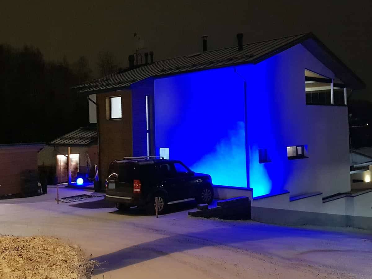 Die Fassade eines modernen Hauses, das nachts von blauen LED-Leuchten beleuchtet wird. Ein Auto im Hof und eine leichte Schneedecke auf dem Boden.