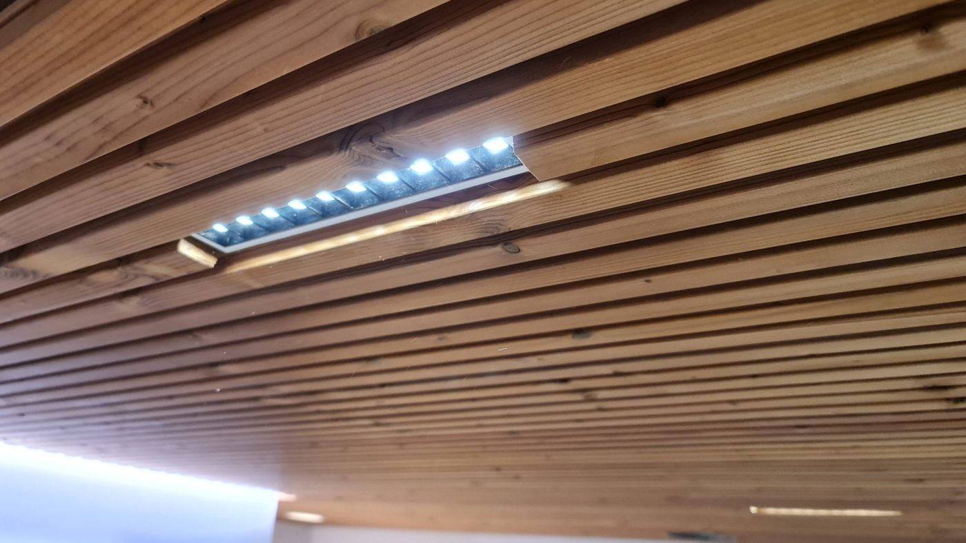 Die Holzdecke mit parallelen Lamellen ist mit einer eingebauten LED-Beleuchtung ausgestattet. Die LEDs sind teilweise durch eine rechteckige Öffnung sichtbar und erzeugen ein subtiles Leuchten auf der natürlichen Holzoberfläche.